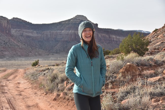 Woman wearing warm hat and coat smiling and standing in desert landscape with redrock cliffs in background.
