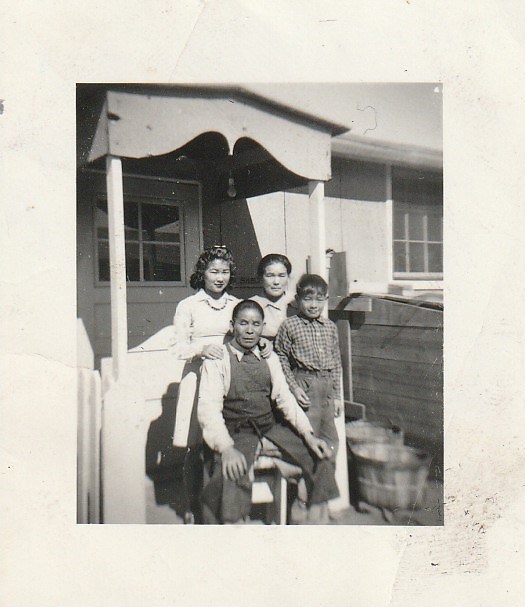 A family of four standing on a front walkway to a house. An older man is sitting with his young son to his right. Behind the father is a young daughter and to her right is her mother.