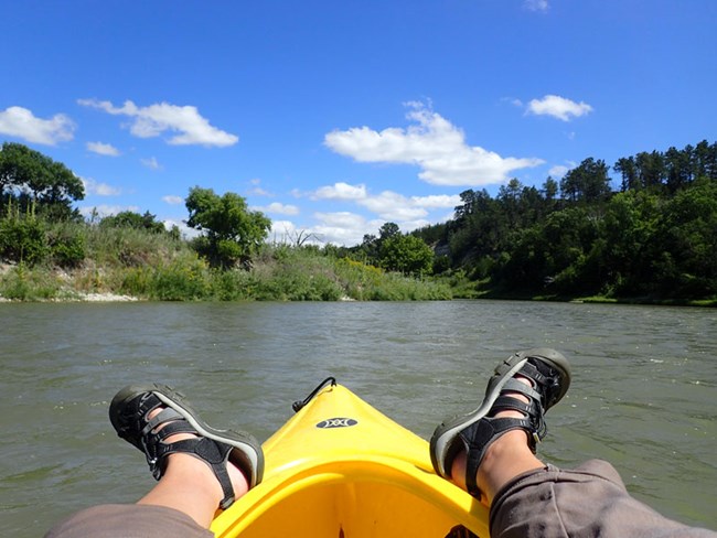 Two legs propped up and over the front of a kayak floating on a calm river.