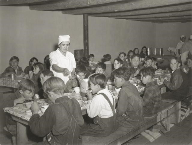 Schoolchildren eating hot school lunches made up primarily of food from the surplus commodities program. Taken at a school in Penasco, New Mexico, United States, December 1941. Photo by the United States Department of Agriculture, public domain. Black and white photo of young kids eating at long lunch tables. A lunch lady in a white smock and hair-covering gestures toward one of the kids. They are in windowless room with a low ceiling and exposed beams.