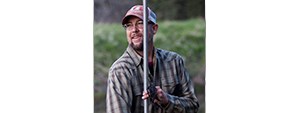 Rob outdoors in a ball cap and plaid shirt, holding up a metal pole.