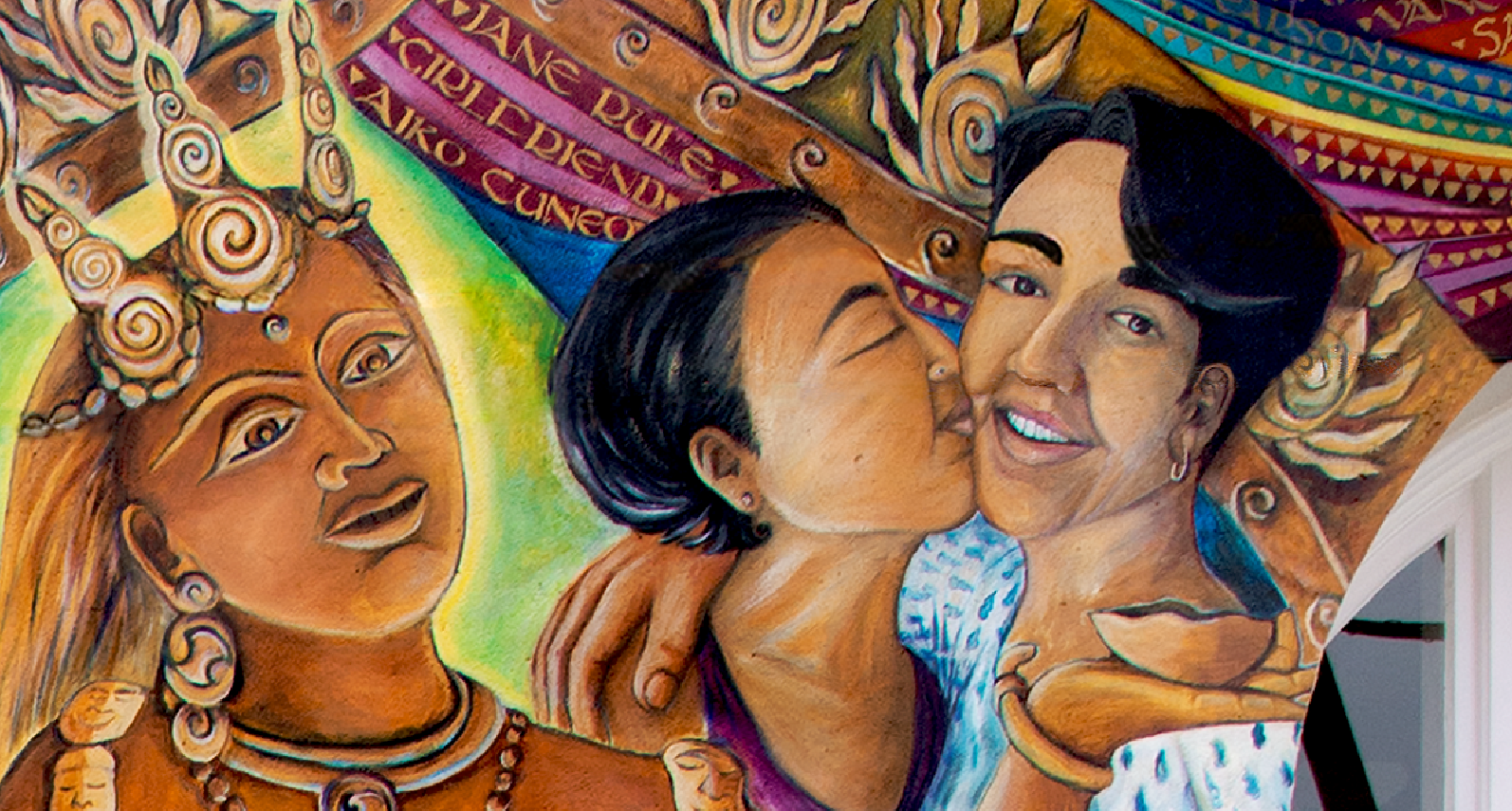 Detail of mural featuring two dark-haired, women embracing. The woman on the right is smiling and the woman on the left kisses her on the cheek. To their left is a South Asian deity wearing a three-pronged crown and ornamental earrings and jewelry.