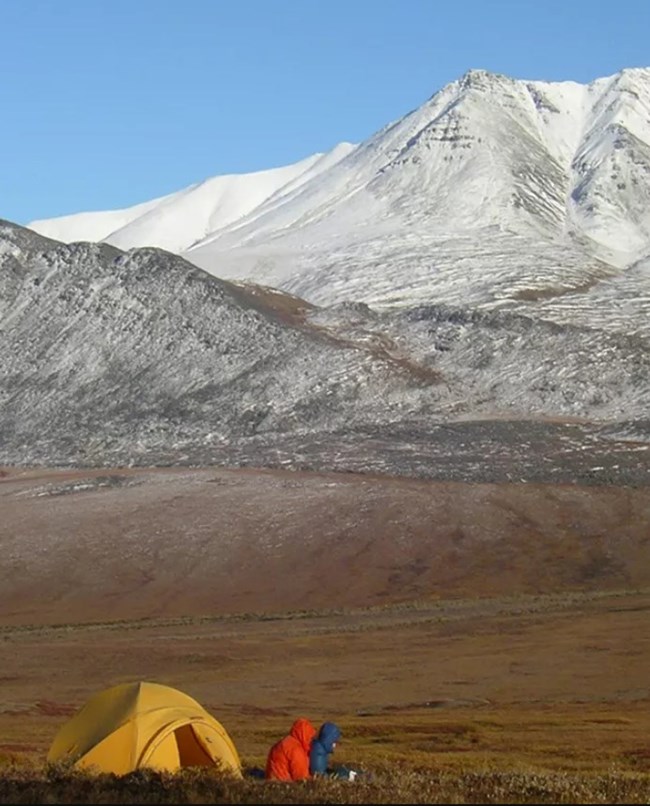two people in warm winter coats sit outside of a yellow tent in a grassy valley with enormous snowy mountains in the distance.
