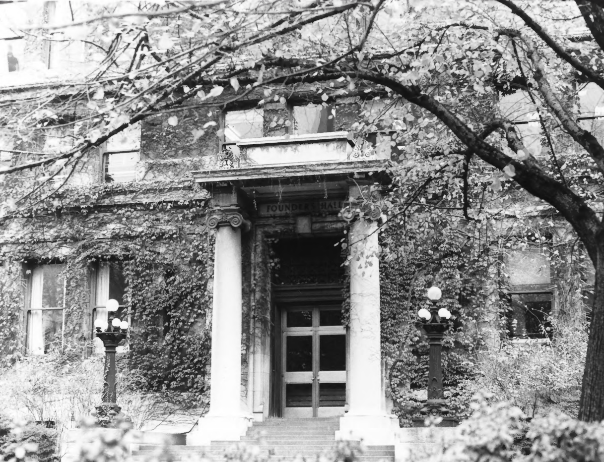 Two Greek columns flank front entrance of ivy-covered, brick, Classical Revival-style building. Low concrete steps lead to door. Tree in bloom cascades over right side. Bushes visible next to short lampposts on either side of entrance.