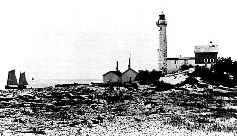 South Manitou Lighthouse Complex, 1883 A grainy black and white photograph with sand and vegetation in the foreground. There are two sailboats in the water on the left. On the right are two small buildings with stacks, and a lighthouse with a long structure attaching it to a small building.