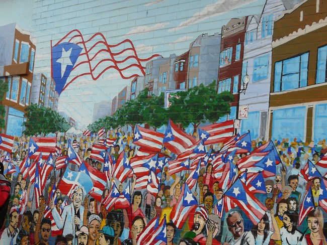 Portion of a mural. A crowd of community members pack Division Street. Most of them are holding Puerto Rican flags. One of the Paseo Boricua Gateway Flags is visible arching over the street.