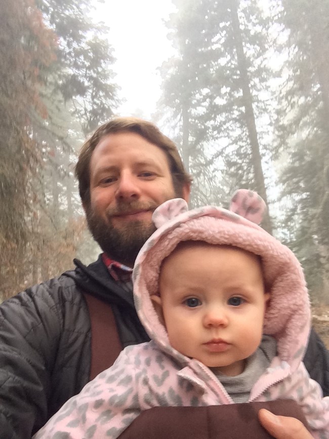 Sean and daughter Man holds his toddler in front of him in the forest.