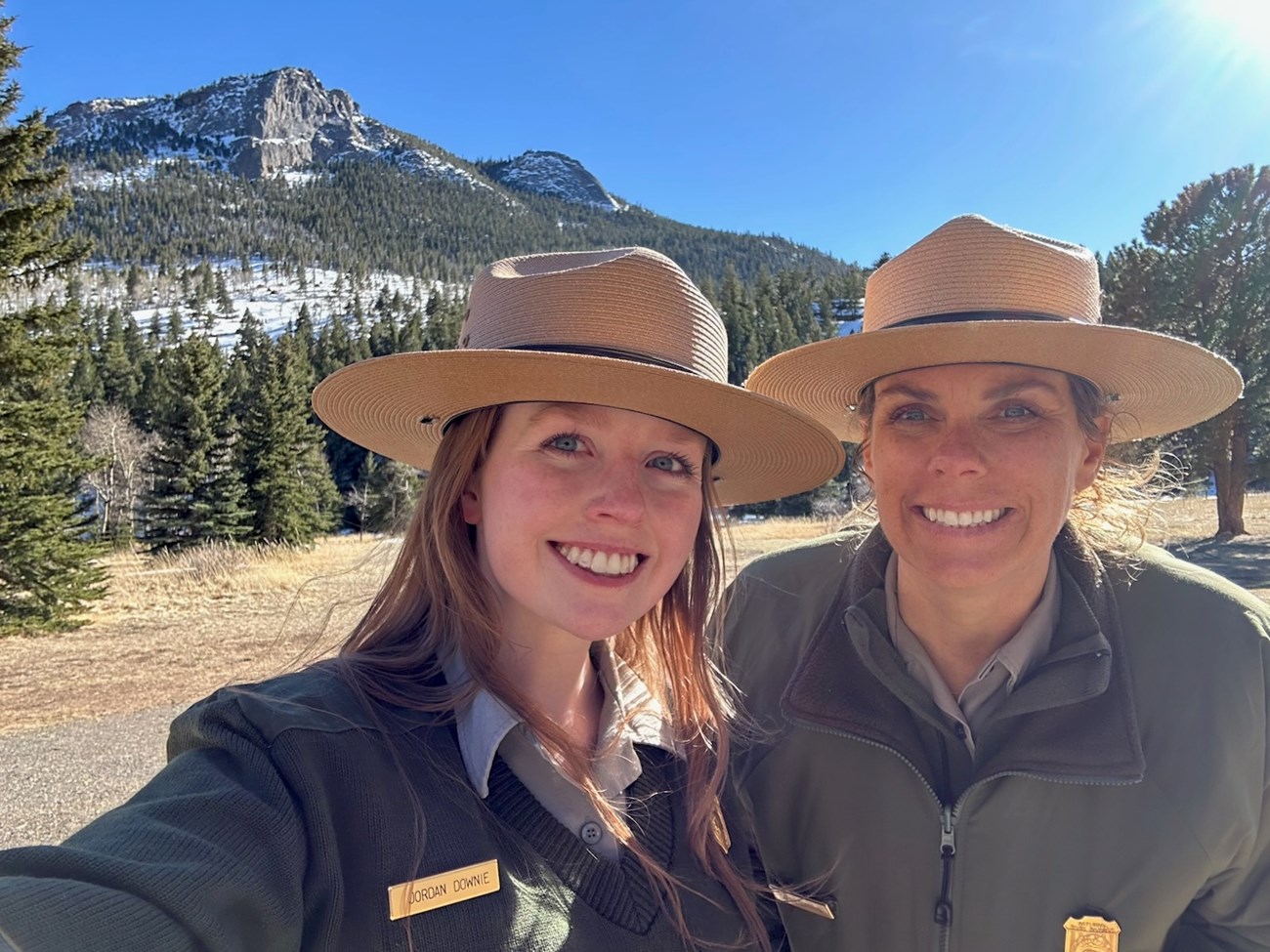 Park rangers and Volunteer Management professionals, Jordan Downie and Sarah Corning pose in Rocky Mountain National Park. Park rangers Jordan Downie and Sarah Corning smile wearing their National Park Service uniforms with a blue sky and mountains behind