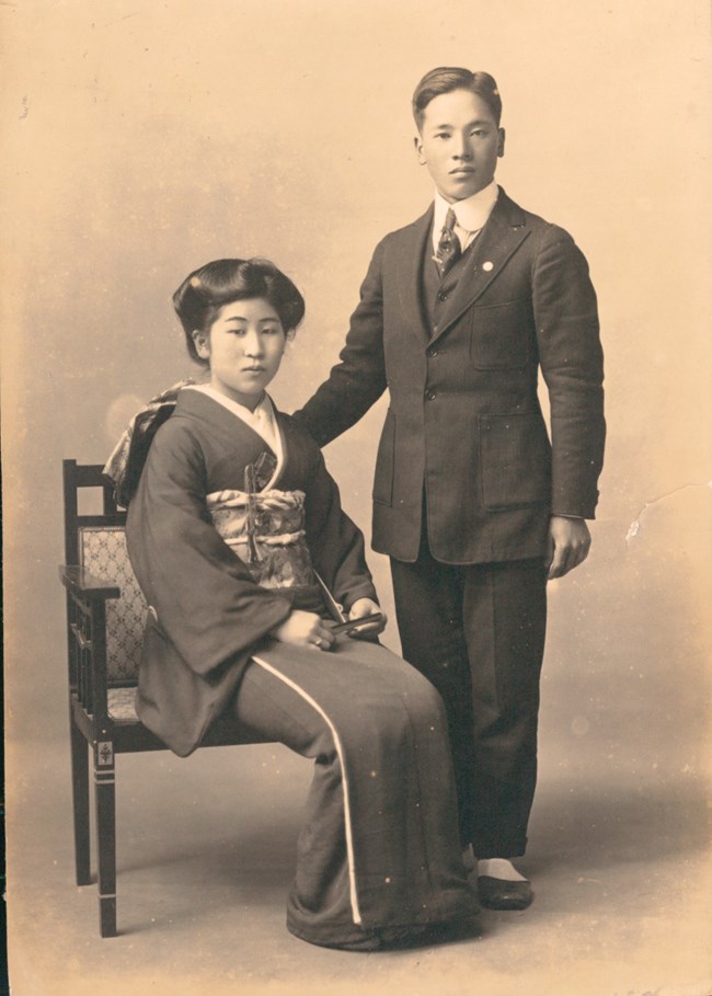 Black and white photo of a Japanese American couple. The woman is seated and wears a traditional Japanese kimono. The man is wearing a western-style suit. He rests his hand on the back of the chair.