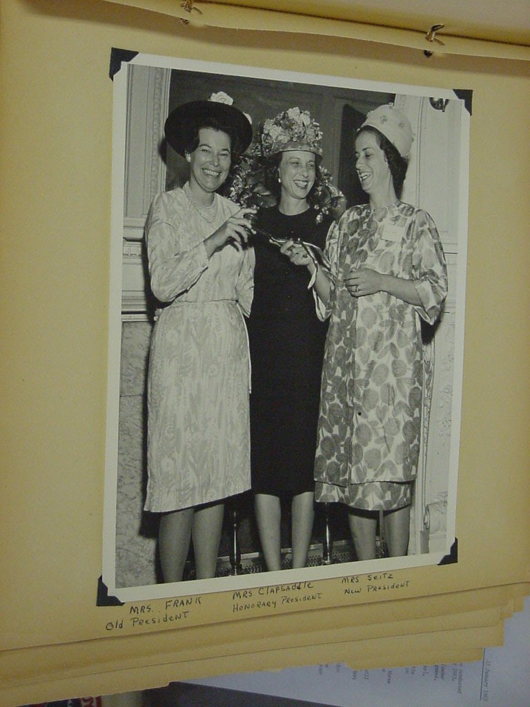 three women in fancy dresses and hats smile and hold a gavel