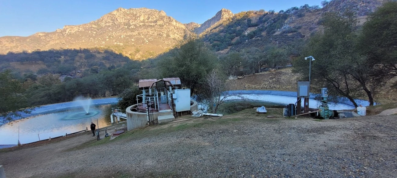 Ash Mountain Wastewater Treatment Plant An out-of-date water pump system sits in front of a man-made water basin with black lining. A mountain range sits behind it.