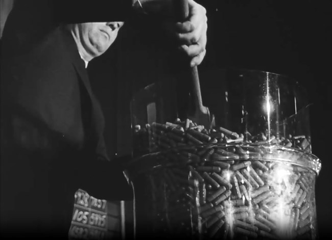 A white man in a dark suit is stirring capsules in a large, clear glass bowl. Behind him you can see a chalkboard with a list of draft numbers already drawn.