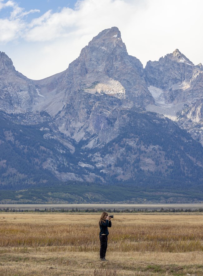 A person stands in a field holding a camera. In the background a large mountain range (the Tetons).