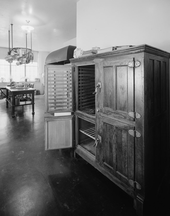 The wooden icebox is located against a wall. One of the paneled doors is open, revealing shelving. In the background is a kitchen work table.