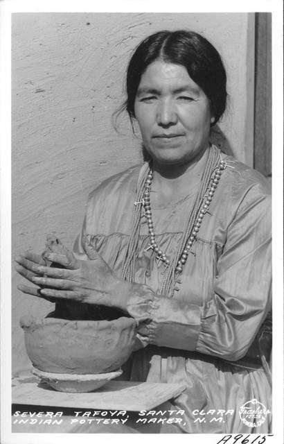 Black and white photo of a woman staring at the camera, shaping clay between her hands with a pot in front of her