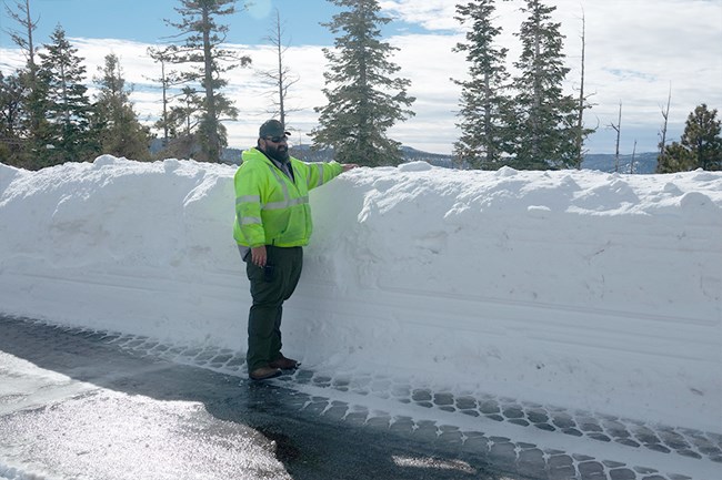 A man in a bright yellow jacket stands beside a tall wall of snow along a road.