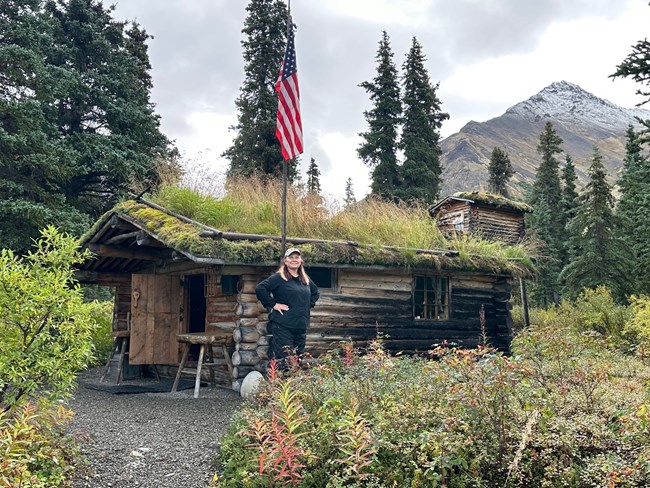 Shelley Curto 1 A person wearing a black shirt and black pants stands in front of a log cabin with a sod roof. There is an American flag on a wooden pole, a mountain, and trees surrounding the cabin.
