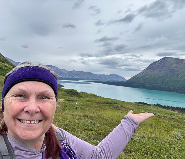 Shelley Curto 4 A smiling woman wearing a purple headband, purple shirt, and carrying a backpack extends an arm toward a view overlooking a blue lake surrounded by green mountains.