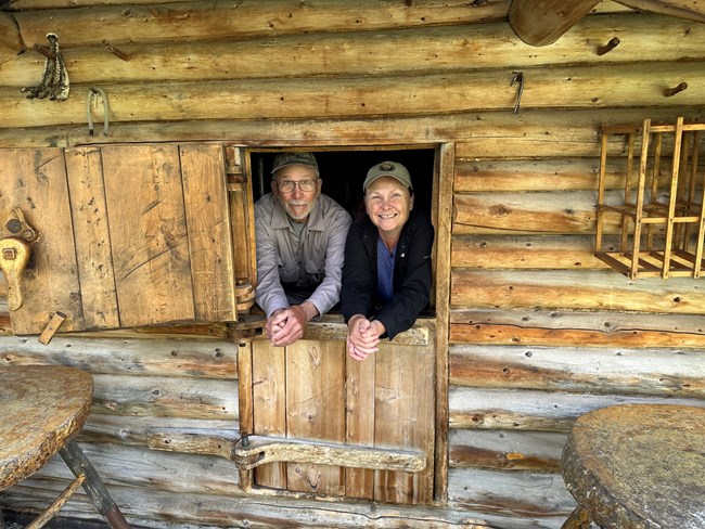 Shelley Curto 7 Two people stand in the doorway of a log cabin. The door is a Dutch door with the bottom half closed and top half open.