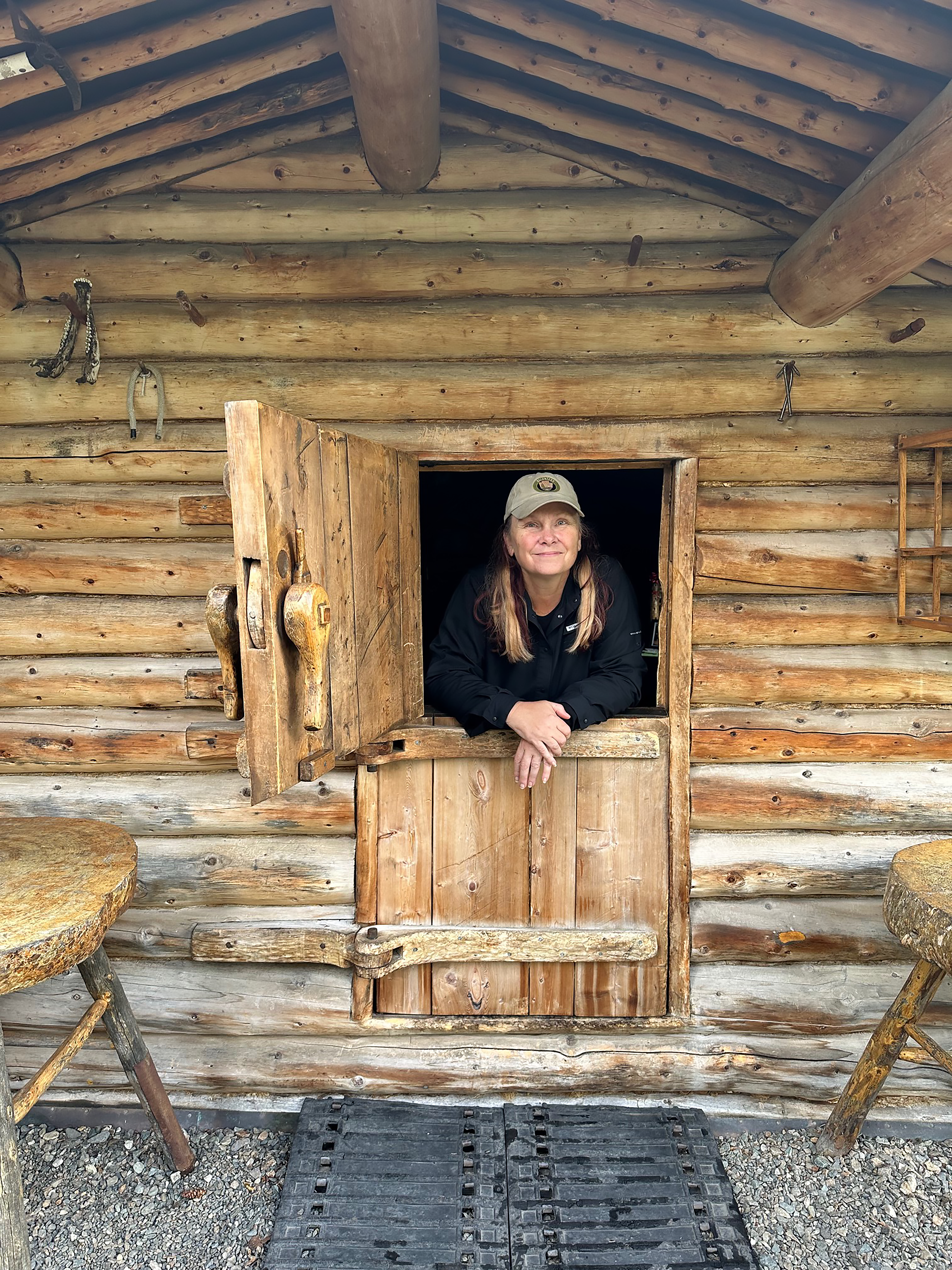 Shelley Curto Proenneke Cabin2 A woman stands in the doorway of a log cabin. The door is a Dutch door with the bottom half closed and the top half open.