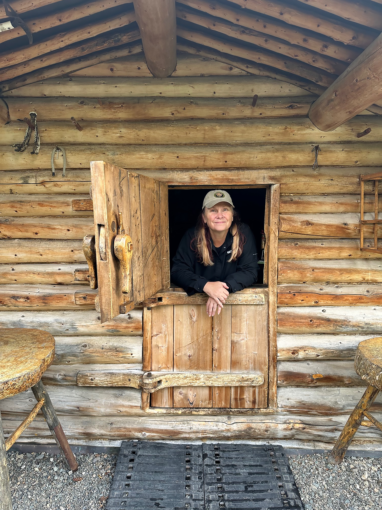A woman stands in the doorway of a log cabin. The door is a Dutch door with the bottom half closed and the top half open.