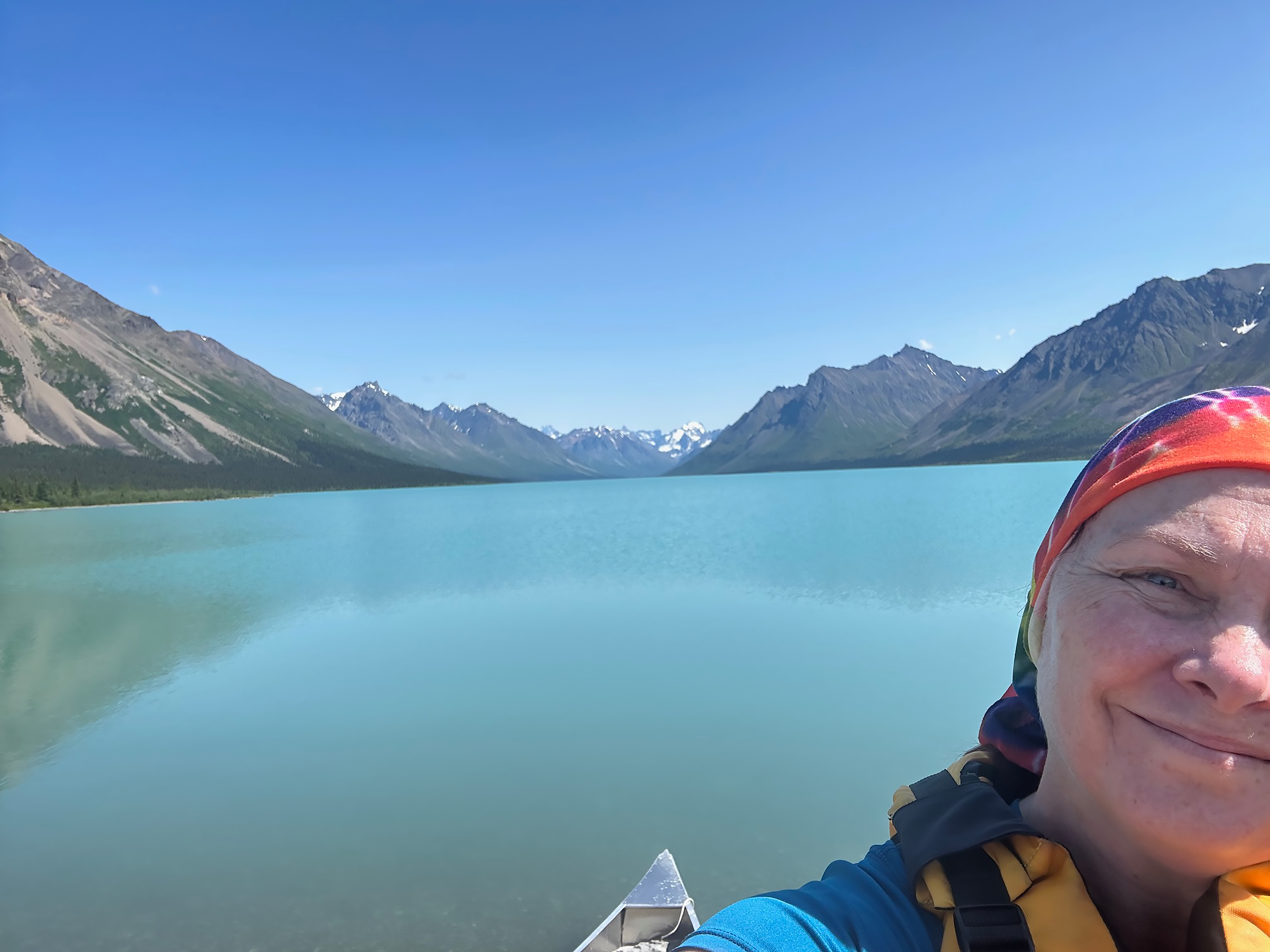Shelley Curto kayak A person wearing a sun protection bandana over their hair, sits in a canoe on a blue lake surrounded by green moutnains.