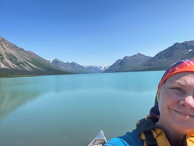 A person wearing a sun protection bandana over their hair, sits in a canoe on a blue lake surrounded by green moutnains.