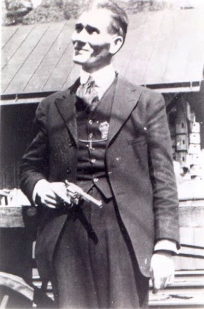William Sidney Hatfield A man in a formal suit with swept back hair stands erect with his head tilted upwards towards the sky and a revolver in his right hand.