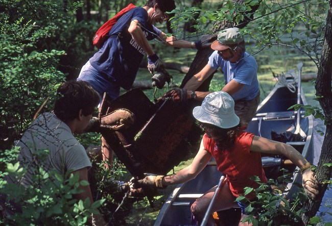 Four people, two in a canoe and two on land, lift a large rusted hunk of metal from a marsh surrounded by green trees and shrubs.
