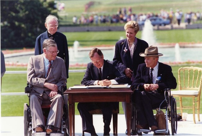 President Bush signs the law while sitting outside at a wooden desk. On either side of the desk are two men in wheelchairs. Behind them stands a man and woman. All four are smiling watching Bush.