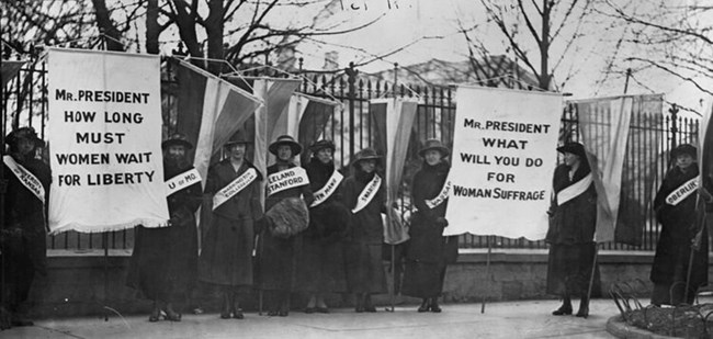 A group of women holding women's suffrage banners stand outside the white house gates. LOC