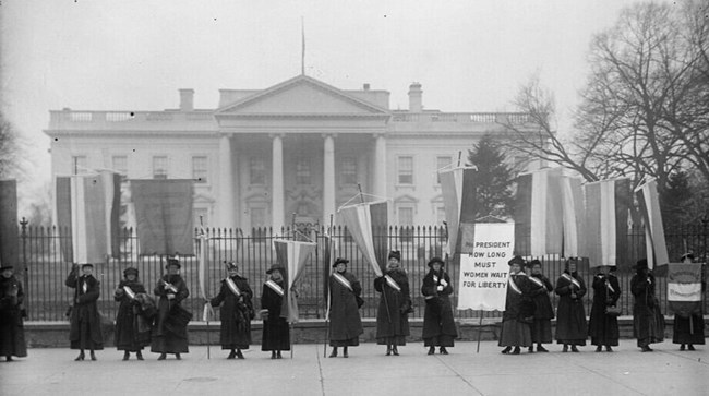 Black and white photo of women in long winter coats standing outside the white house fence LOC