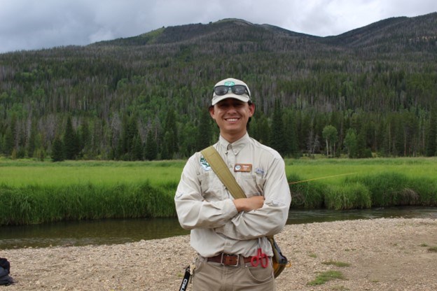 Simon Aceto outside A young individual posing in front of green lush mountains, wearing a hat, with arms crossed.