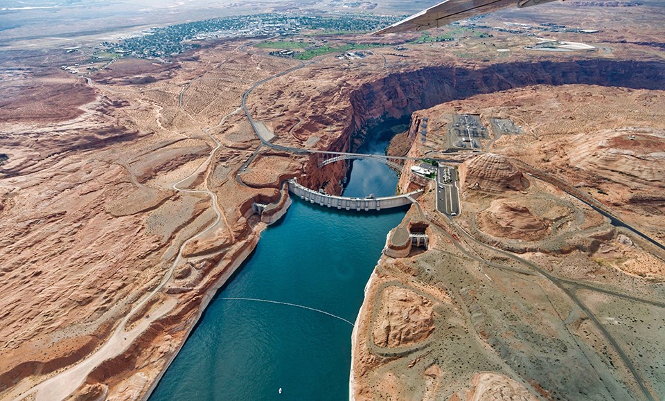 Glen Canyon Dam Slick rock Landscape with broad blue river blocked by semi-circular concrete structure. A bridge spans the canyon after the concrete structure.
