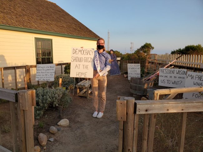 Photo of Sita Antel standing in the “picket garden” she created holding a sign that reads “Democracy should begin at the home.”