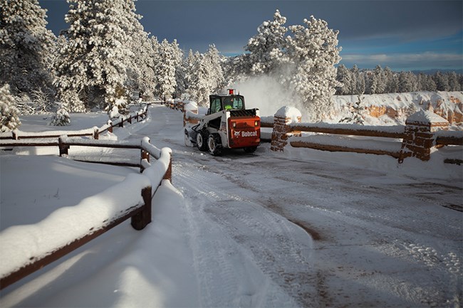 A skid-steer tractor snow blows snow along a sidewalk