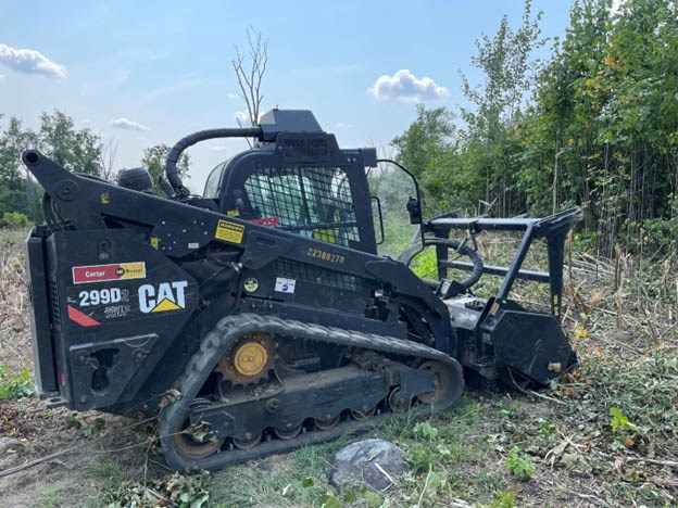 Small construction machine with treads and a front mower type attachment sits in a field.
