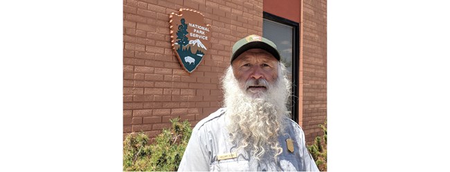 A man in an NPS uniform with a long white beard stands in front of a red brick building with an NPS logo.