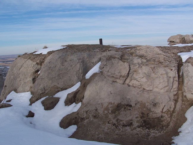 A metal post sticks out of a large section of sandstone that is partially snow covered.