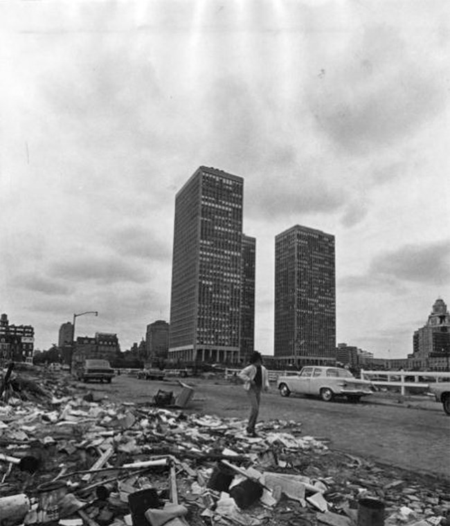 A man walking through the rubble of demolished buildings with new apartment buildings behind him.