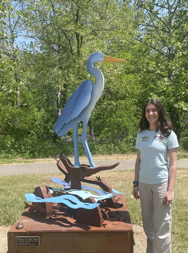 A woman with brown hair wearing a blue uniform shirt stands next to a metal statue of a great blue heron.