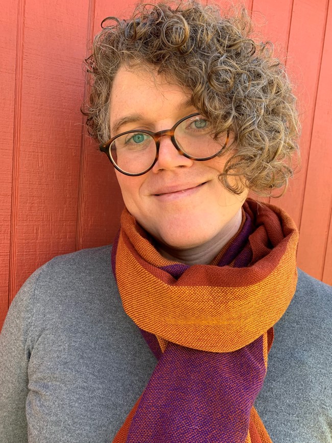 Individual with curly hair and glasses stands in front of a red background