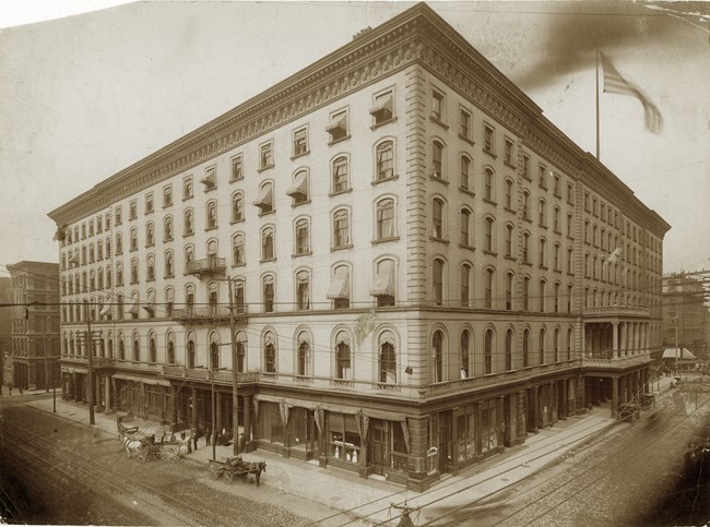 Southern Hotel Rebuilt after 1877 Fire - MOHIST Sepia colored photo of a six-story square building with an American flag in the top right corner.