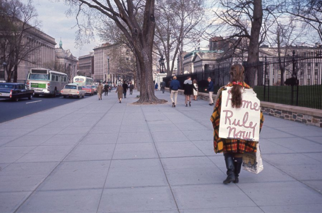 A person stands on a sidewalk, wearing a sandwhich board sign that says "home rule now"