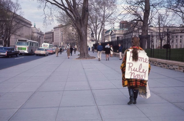 Woman protesting for Home Rule outside of White House. 1966. A person stands on a sidewalk, wearing a sandwhich board sign that says "home rule now"