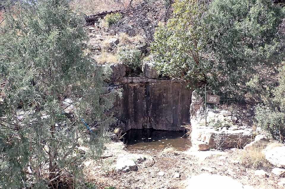 Dog Canyon Spring A spring bounded by three carved vertical rock walls. A fence on the perimeter has a sign that reads “FRAGILE SPRING KEEP OUT”.