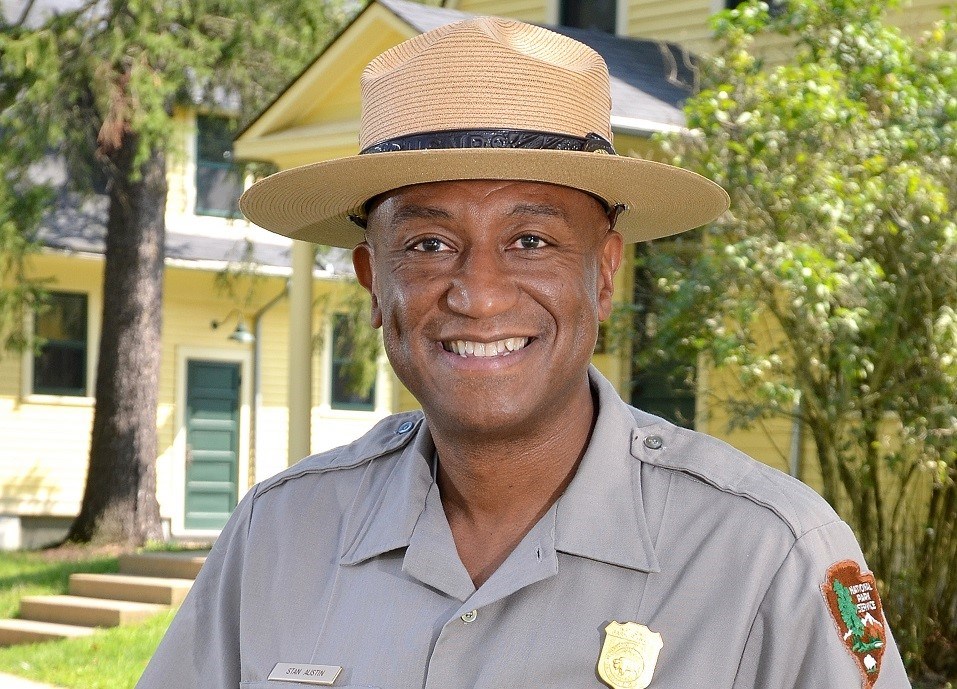 Smiling man with dark skin and broad-brimmed hat stands in front of a large yellow house with green door.