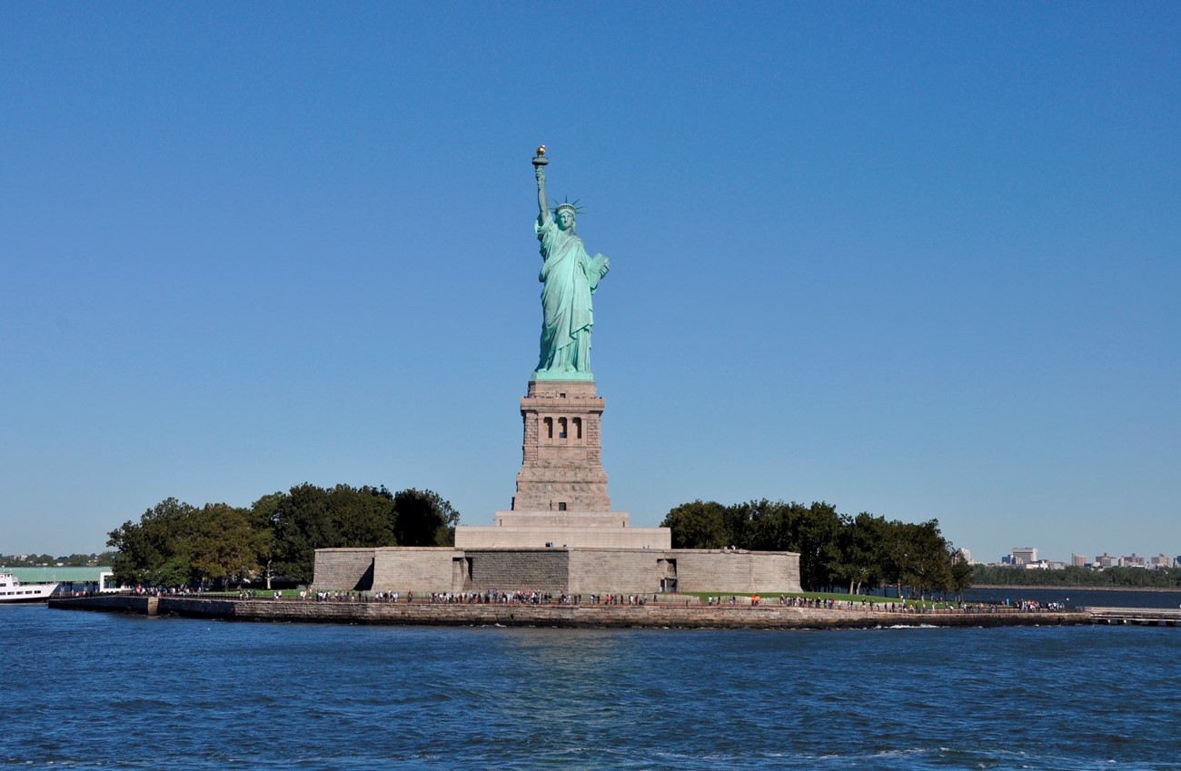 Green-patina covered copper colossal statue of a woman with a crown and uplifted right arm stands on a granite pedestal on an island.