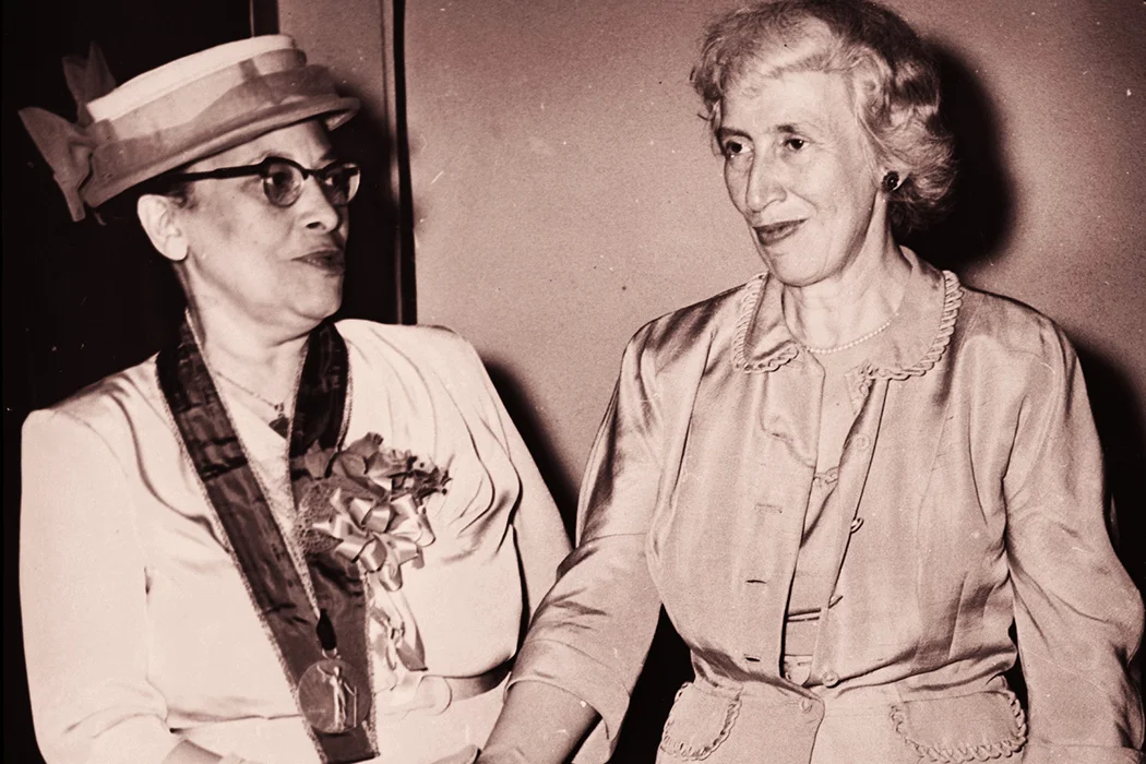 Black and white photo of two women. The Black woman on left is wearing a hat with a bow, cat-eye glasses, a corsage and a medal. She is shaking the hand of the white woman on the right in a collared dress and round earrings.