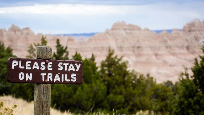 a sign that says "please stay on trails" with badlands hills and trees in the background